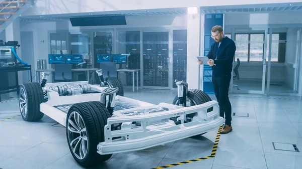 Engenheiro com óculos e barba verifica o protótipo do chassi do carro elétrico com rodas, baterias e motor com um software de realidade aumentada em um computador tablet em um laboratório de desenvolvimento de alta tecnologia . — Fotografia de Stock