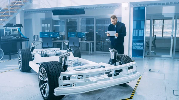 Ingeniero con gafas y barba escanea el prototipo de chasis de coche eléctrico con ruedas, baterías y motor con un software de realidad aumentada en una tableta en un laboratorio de desarrollo de alta tecnología . —  Fotos de Stock