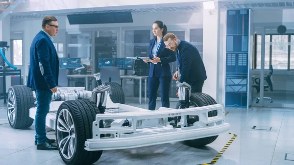 Automotive design ingenjörer pratar medan du arbetar på elbil chassi prototyp. I innovation laboratorie anläggning koncept fordons ram inkluderar hjul, fjädring, motor och batteri. — Stockfoto