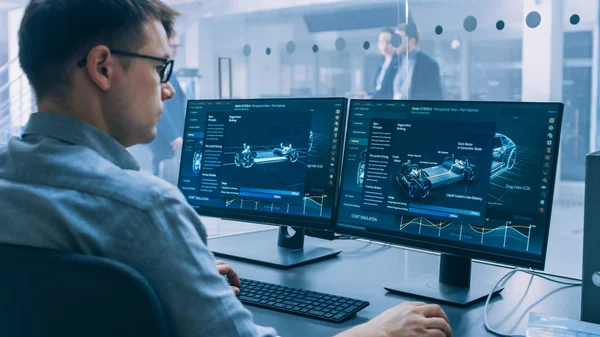 Professioneller Ingenieur arbeitet an einem Computer mit 3D-Cad-Software und testet den Prototyp eines Elektroauto-Fahrgestells mit Rädern, Batterien und Motor stehend in einem Hightech-Entwicklungslabor. — Stockfoto