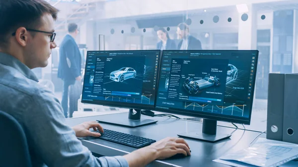 Professional Engineer Works on a Computer with a 3D CAD Software and Tests the Electric Car Chassis Prototype with Wheels, Batteries and Engine Standing in a High Tech Development Laboratory. — Stock Photo, Image