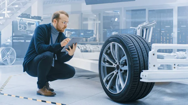 Ingeniero con gafas y barba trabaja en una tableta al lado de un prototipo de chasis eléctrico con ruedas, baterías y motor en un laboratorio de desarrollo de alta tecnología . — Foto de Stock