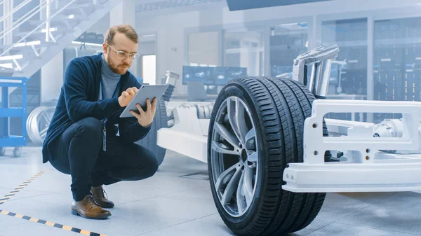 Engenheiro com óculos e barba funciona em um computador tablet ao lado de um protótipo de chassi de carro elétrico com rodas, baterias e motor em um laboratório de desenvolvimento de alta tecnologia . — Fotografia de Stock