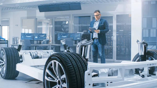 Automotive design ingenjör tittar på teknisk ritning av en elbil chassi prototyp på en tablett. I innovation laboratorie anläggning koncept fordons ram innehåller hjul, motor och batteri. — Stockfoto