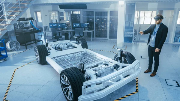 증강 현실 헤드셋을 사용하여 전기 자동차 섀시 플랫폼에서 작업하는 자동차 엔지니어. 혁신 실험실 시설 개념 차량 프레임에는 바퀴, 서스펜션, 엔진 및 배터리가 포함됩니다.. — 스톡 사진