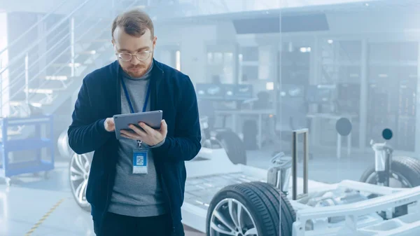 Homme ingénieur travaille dans une installation de développement de haute technologie détenant un ordinateur tablette. Il se tient à côté d'un prototype de châssis de voiture électrique avec piles et roues . — Photo