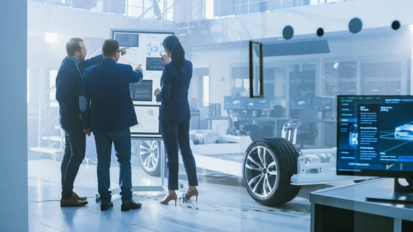 Diverse Team of Design Engineers are Working and Discussing Something on a Interactive Whiteboard Next to an Electric Car Chassis Prototype. In High Tech Laboratory Facility with Vehicle Frame. — Stock Photo, Image