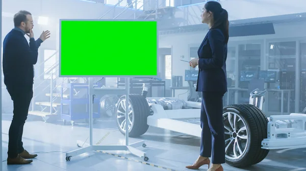 Ingenieros de diseño masculino y femenino están discutiendo algo en una pizarra interactiva con pantalla verde junto a un prototipo de chasis de coche eléctrico. En las instalaciones de laboratorio de alta tecnología con el marco del vehículo . — Foto de Stock