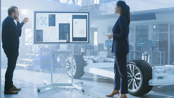 Mężczyzna i kobieta Design Engineers pracują i omawiają coś na tablicy interaktywnej obok samochodu elektrycznego prototyp podwozia. W zakładzie laboratoryjnym High Tech z ramą pojazdu. — Zdjęcie stockowe