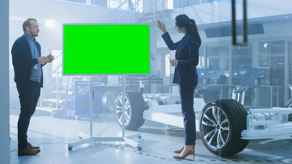 Mannelijke en vrouwelijke ontwerpingenieurs bespreken iets op een interactief whiteboard met groen scherm naast een elektrisch auto chassis prototype. In hightech laboratorium faciliteit met voertuig frame. — Stockfoto