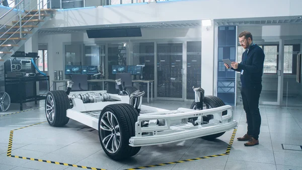 Ingeniero con gafas y barba escanea el prototipo de chasis de coche eléctrico con ruedas, baterías y motor con un software de realidad aumentada en una tableta en un laboratorio de desarrollo de alta tecnología . —  Fotos de Stock