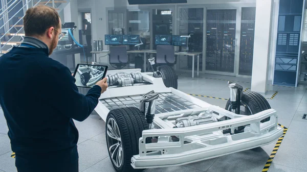 Ingeniero con gafas y barba escanea el prototipo de chasis de coche eléctrico con ruedas, baterías y motor con un software de realidad aumentada en una tableta en un laboratorio de desarrollo de alta tecnología . — Foto de Stock