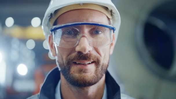 Mosolygós professzionális nehézipari mérnök / munkás biztonsági egyenruha, szemüveg és keménykalap portréja. A háttérben fókuszálatlan nagy ipari gyár — Stock videók