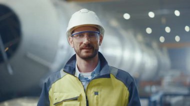 Gülümseyen Profesyonel Ağır Sanayi Mühendisi / İşçi Güvenlik Üniforması, Gözlük ve Sert Şapka Giyen Portresi. Kaynağın kıvılcımlar saçtığı Odaklanmamış Büyük Endüstriyel Fabrikada