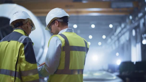 Twee zware industrie ingenieurs staan in Pipe Manufacturing Factory, gebruik maken van digitale tablet computer, hebben discussie. Grote pijp in elkaar gezet. Ontwerp en bouw van een transportleiding voor olie, gas en brandstoffen — Stockvideo