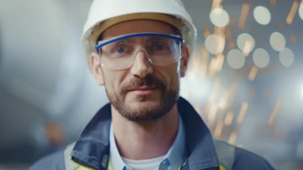 Porträtt av Leende Professionell Tung Industri Ingenjör / Arbetare Bär Säkerhet Uniform, Glasögon och Hård Hatt. I bakgrunden ofokuserad stor industrifabrik där svetsgnistor flyger — Stockvideo