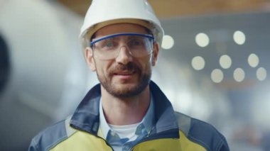 Profesyonel Ağır Sanayi Mühendisi / İşçi Güvenlik Üniforması, Gözlük ve Sert Şapka Gülümseyen Portresi. Kaynağın kıvılcımlar saçtığı Odaklanmamış Büyük Endüstriyel Fabrikada