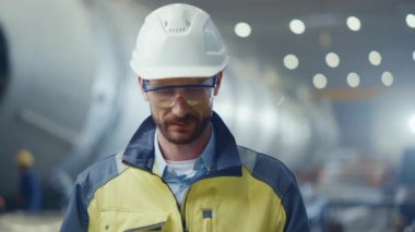 Profesyonel Ağır Sanayi Mühendisi / İşçi Güvenlik Üniforması, Gözlük ve Sert Şapka Gülümseyen Portresi. Kaynağın kıvılcımlar saçtığı Odaklanmamış Büyük Endüstriyel Fabrikada