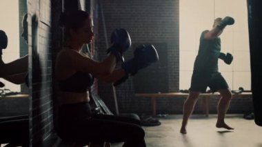 Güzel Fit Esmer Kickboxer Atlet Motivasyonposterler ile Loft Gym bir Bankta Oturan. Mavi Dövüş Eldivenlerini Takıyor. Kadın Ayağa Kalkar ve Onun Yoğun Boks Eğitimi Devam Ediyor.