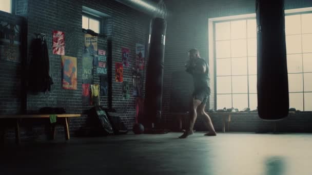 Hombre fuerte y guapo Kickboxer está trabajando en un gimnasio Loft con carteles motivacionales en las paredes. El atleta masculino está practicando golpes en una bolsa de boxeo. Es serio y enérgico. . — Vídeos de Stock