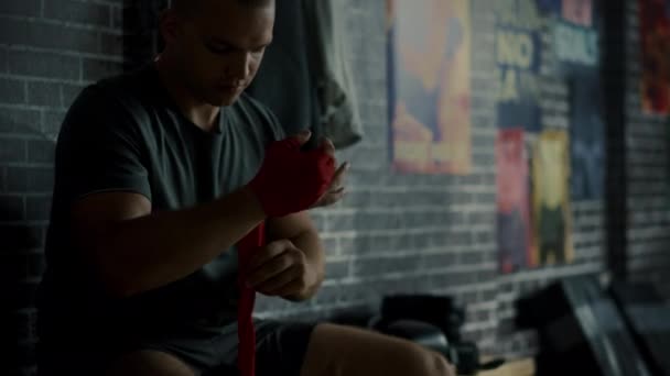 Silny mężczyzna zawijania ręce z Handwrapping rozpocząć jego kickboxing Workout w siłowni z motywacyjne plakaty na ścianach. Używa czerwone pasy wokół nadgarstka, kciuka i Knuckles. Strzał z bliska. — Wideo stockowe