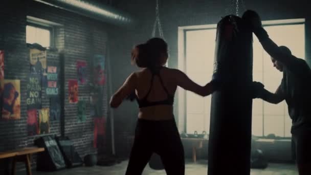 Fit атлетична жінка Трамбором удари і показів боксерської груші під час тренування в лофт тренажерний зал. Вона прекрасна і енергійна. Сильний тренер тримає боксерський мішок. Тренінг «інтенсивна самозахист». — стокове відео