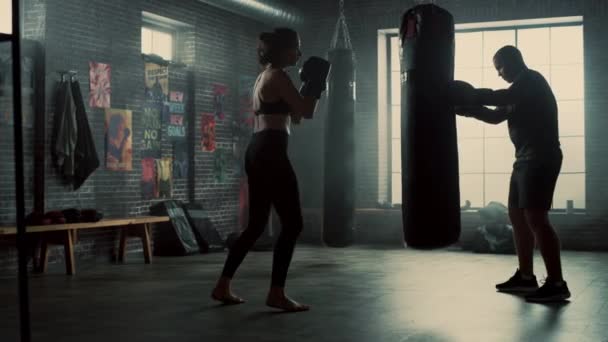 Fit atletisk kvinna Kickboxer stansar och träffar slagsäck under ett träningspass i ett loft gym. Hon är vacker och energisk. Strong Trainer håller boxnings väskan. Intensiv självförsvars utbildning. — Stockvideo