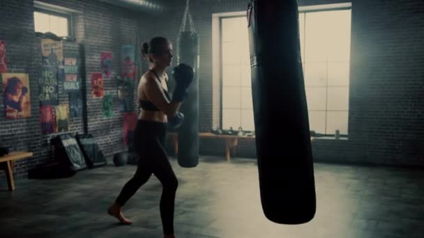 Calzata atletica donna Kickboxer pugni e colpisce il sacco da boxe durante un allenamento in una palestra Loft con poster motivazionali. E 'esausta e stanca dopo il suo intenso addestramento di autodifesa. . — Video Stock