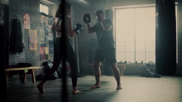 フィットアスレチック女性キックボクサーパンチとロフトジムでのワークアウト中にパンチパッドを打ちます。彼女は美しくてエネルギッシュです。強いコーチはボクシングパッドを保持しています。激しい自衛訓練. — ストック動画