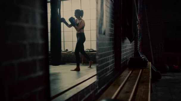 Fit Athletic Woman Kickboxer golpea y golpea el saco de boxeo durante un entrenamiento en un gimnasio loft con carteles motivacionales. Está agotada y cansada después de su intenso entrenamiento de autodefensa. Disparo en un espejo . — Vídeo de stock