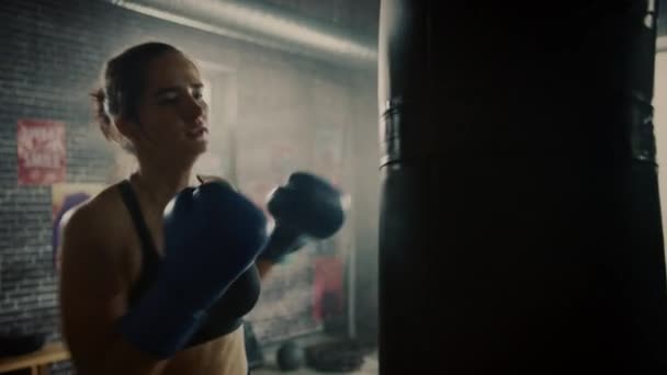 Die fitte athletische Kickboxerin schlägt beim Training in einer Loft-Turnhalle mit Motivationsplakaten auf den Boxsack ein. Sie ist erschöpft und müde nach ihrem intensiven Selbstverteidigungstraining. — Stockvideo