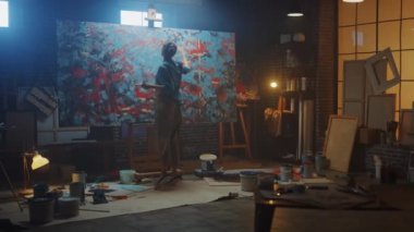 Yetenekli Kadın Sanatçı Bir Tabloya Yürüyor ve Modern Soyut Yağlı Boyama Boyası Üzerinde Çalışmaya Başlıyor. Karanlık Yaratıcı Stüdyo Büyük Resim Easel Aydınlanmış, Araçlar, Her Yerde Boyama Üzerine Duruyor
