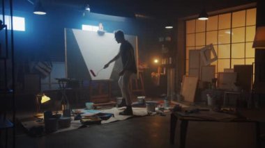 Yetenekli erkek ressam soyut bir resim üzerinde çalışıyor, Endüstriyel Roller 'ı kullanarak cesur duygusal bir modern resim çiziyor. Easel Aydınlatma Üzerine Karanlık Yaratıcı Stüdyo Büyük Tuval Standı. Yavaş Hareket