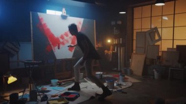 Yetenekli erkek ressam soyut bir resim üzerinde çalışıyor, Endüstriyel Roller 'ı kullanarak cesur duygusal bir modern resim çiziyor. Easel Aydınlatma Üzerine Karanlık Yaratıcı Stüdyo Büyük Tuval Standı. Yavaş Hareket