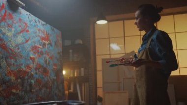 Yetenekli Kadın Sanatçı Enerjik ve Profesyonel Boya Fırçası Kullanarak O Yağlıboya Modern Masterpiece oluşturur. Çarpıcı Renkler Büyük Tuval ile Yaratıcı Stüdyo. Düşük Açılı Yan Görünüm