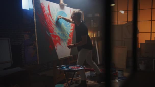 Talentierter Künstler, der an abstrakter Malerei arbeitet, verwendet Pinsel, um ein kühn emotionales modernes Bild zu schaffen. dunkles Kreativatelier große Leinwand steht auf Staffelei beleuchtet. Seitenansicht Bogenschuss — Stockvideo