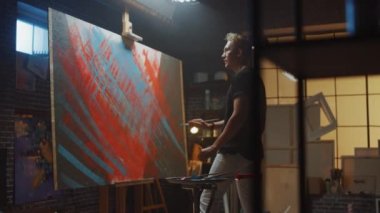 Yetenekli erkek Sanatçı Soyut Yağlıboya Üzerine Çalışır, Boya Fırçageniş vuruş ile o Modern Başyapıt oluşturur. Koyu ve Dağınık Yaratıcı Stüdyo nerede Büyük Tuval Easel Işıklı Standları