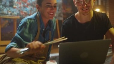 Stüdyolarında Oturan Birkaç Sanatçı Dizüstü Bilgisayar Kullanıyor, Bir Proje Üzerinde Çalışıyor, Konuşuyor ve Gülümsüyor. Ressam ve Ortağı Araştırma Yapıyor. Yağlıboya Resim ve Aletlerle Otantik Atölye