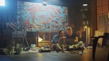 Stüdyolarında Oturan Birkaç Sanatçı Dizüstü Bilgisayar Kullanıyor, Bir Proje Üzerinde Çalışıyor, Konuşuyor ve Gülümsüyor. Ressam ve Ortağı Araştırma Yapıyor. Yağlıboya Resim ve Aletlerle Otantik Atölye