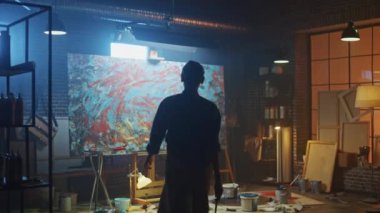 Kendine güvenen ve Kararlı Kadın Sanatçı Tuval için Walks ve Paintbrush kullanarak Modern Soyut Yağlıboya Çalışmaya Başlar. Yaratıcı Studio Büyük Resim Easel Işıklı duruyor. Geri Görünüm Çekimini Takip Edin