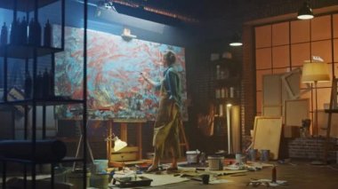 Kendine güvenen ve Kararlı Kadın Sanatçı Tuval için Walks ve Paintbrush kullanarak Modern Soyut Yağlıboya Çalışmaya Başlar. Yaratıcı Studio Büyük Resim Easel Işıklı duruyor. Ark Arka Görünüm Çekimi