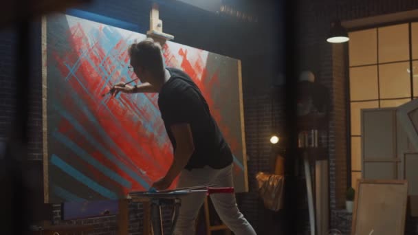 Talented male Artist Works on Abstract Oil Painting, with Broad Strokes of Paint Brush he Creates Modern Masterpiece. Dark and Messy Creative Studio where Large Canvas Stands on Easel Illuminated — Stock Video