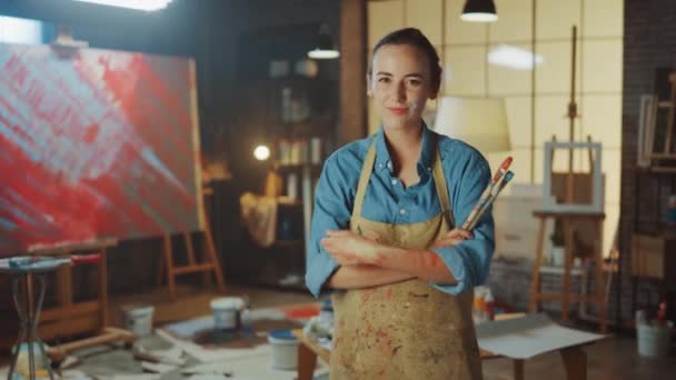 Retrato de talentosa joven artista sucia con pintura, usando delantal y sosteniendo pinceles mira a la cámara sonriendo. Auténtico estudio creativo con grandes lienzos y equipos por ahí — Vídeo de stock