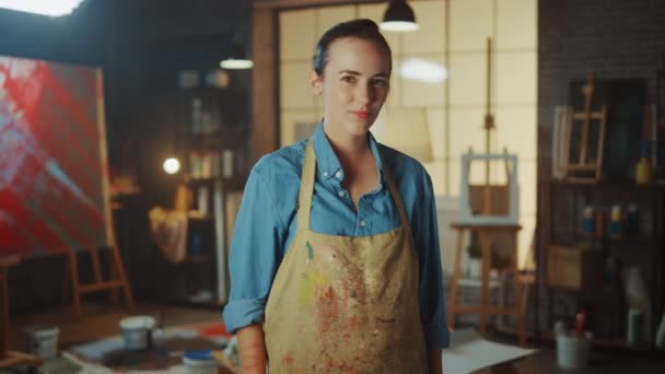 Retrato de talentosa joven artista sucia con pintura, usando delantal, cruza brazos mientras sostiene cepillos, mira a la cámara con una sonrisa. Auténtico Estudio Creativo con Lienzo Grande y Herramientas en Todas Partes — Vídeo de stock