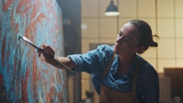 Female Artist Works on Abstract Oil Painting, Moving Paint Brush Energetically She Creates Modern Masterpiece. Dark Creative Studio where Large Canvas Stands on Easel Illuminated. Zoom Out Shot — Stock Video