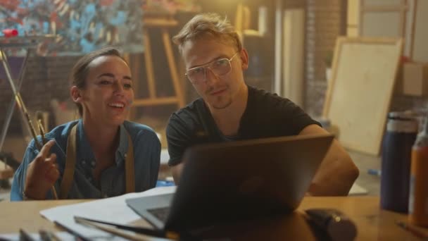 Paar artiesten zitten in hun studio gebruik laptop computer, werken aan een project. Schilder en haar mentor doen onderzoek. In de achtergrond authentieke workshop met olieverfschilderijen en gereedschap — Stockvideo