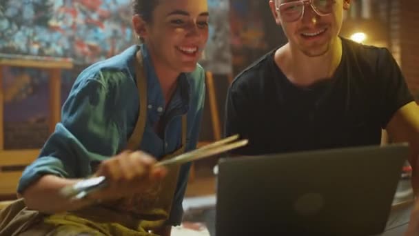 Einige Künstler, die in ihrem Atelier sitzen, benutzen Laptop-Computer, arbeiten an einem Projekt, reden und lächeln. Malerin und ihr Partner bei der Recherche. authentische Werkstatt mit Ölgemälden und Werkzeugen — Stockvideo