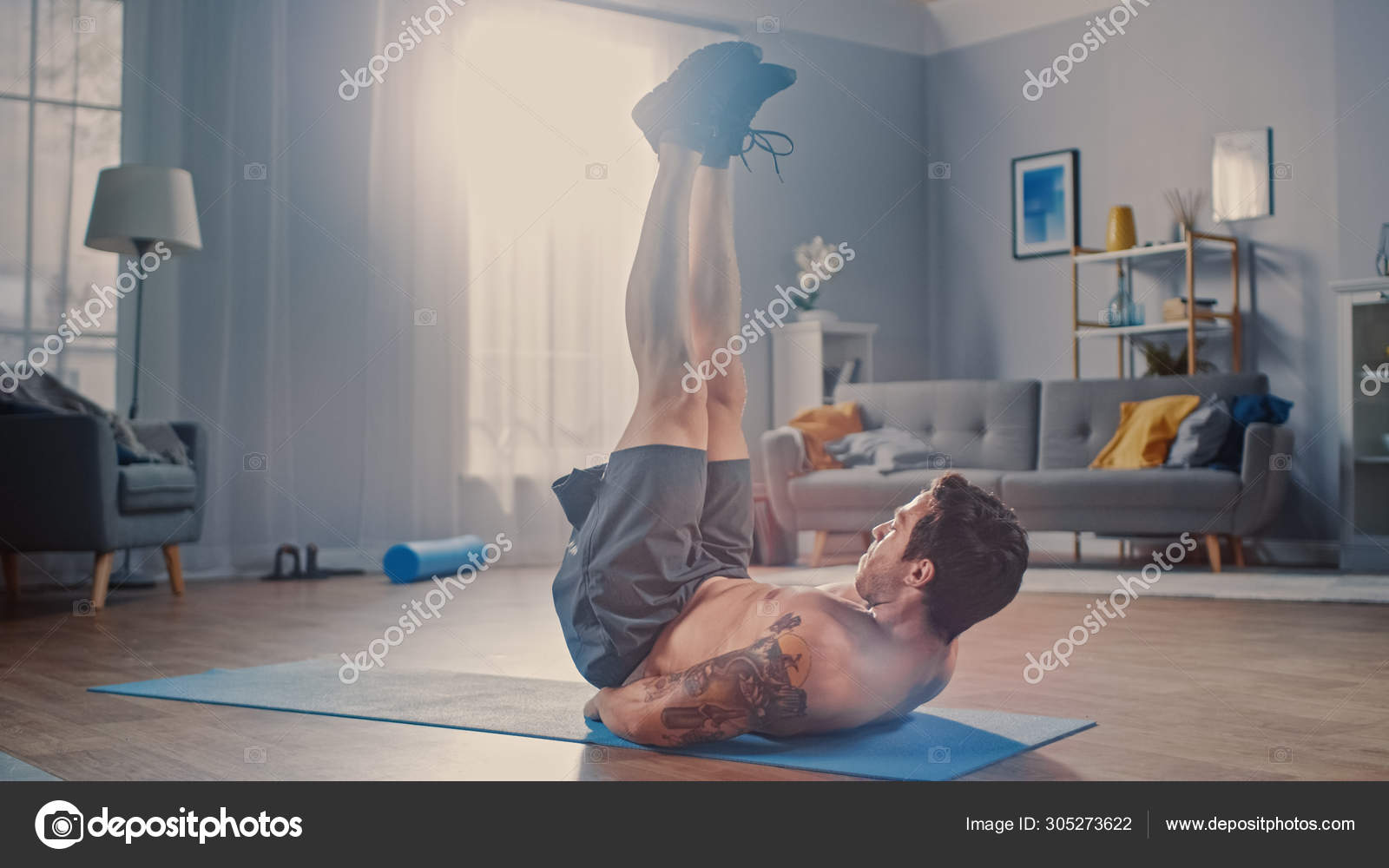 强大的运动无衬衫适合男子在灰色短裤是做腹肌练习在家里在他的宽敞和阳光明媚的客厅与简约的内部 图库照片 C Gorodenkoff