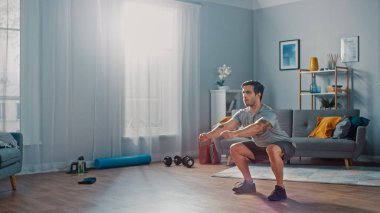 T-shirt ve Şort Kas Athletic Fit Man Minimalist İç ile Onun Geniş ve Parlak Oturma Odasında Evde Squat Egzersizleri yapıyor.