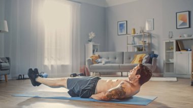 Gri Şort Güçlü Atletik Gömleksiz Fit Man Minimalist İç ile Onun Geniş ve Güneşli Oturma Odasında Evde Karın Egzersizleri yapıyor.